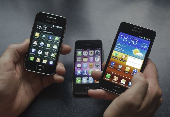 Vyras rankose laiko „Samsung Galaxy S II“ (d.) ir „Galaxy Ace“ išmaniuosius telefonus. Fone – „Apple“ telefonas „iPhone 4“.